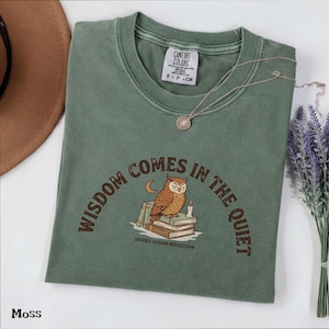 以下が含まれることがあります： モスグリーンのクルーネックTシャツ。本の上に止まるフクロウ、キャンドル、三日月のグラフィックが描かれています。テキストは「WISDOM COMES IN THE QUIET」および「SPOOKY SEASON REFLECTION」と記されています。Comfort Colorsのブランドラベル付き。
