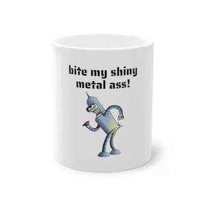 Op de afbeelding: Witte keramische mok met het stripfiguur Bender uit Futurama. De mok toont de zin "bite my shiny metal ass!" in het zwart. Bender is afgebeeld in metallic grijs met een blauwe vonk.