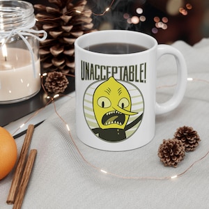 May include: White ceramic mug featuring a cartoon lemon character with the word "UNACCEPTABLE!" above it. The lemon has a yellow face, black eyes, and a long nose. The mug is filled with a dark liquid.