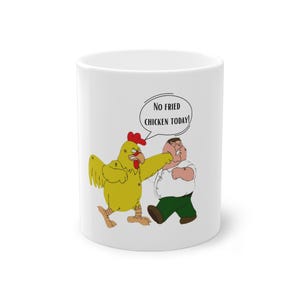 May include: White ceramic mug featuring a cartoon chicken punching a man. The chicken is yellow with a red comb and the man is wearing a white shirt and green pants. Text in a speech bubble reads "No Fried Chicken Today!"