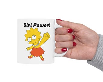 May include: White ceramic mug featuring a cartoon image of Lisa Simpson with the words "Girl Power!". Lisa is depicted with yellow hair, an orange dress, and a pearl necklace. The mug is held by a hand with red nail polish.