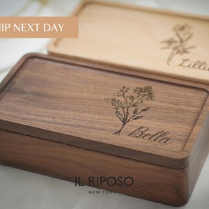 Puede incluir: Dos cajas rectangulares de madera con tapas. Una caja es de color marr&oacute;n oscuro con un dise&ntilde;o floral y el nombre "Bella" grabado en la tapa. La otra caja es de color marr&oacute;n claro con un dise&ntilde;o floral y el nombre "Lillian" grabado. El texto "SHIP NEXT DAY" es visible.