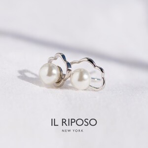 May include: A pair of silver-toned earrings featuring a pearl within a stylized, cloud-shaped frame. The earrings are set against a white background, with the brand name "IL RIPOSO NEW YORK" visible below.