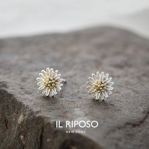 May include: A pair of daisy-shaped stud earrings. Each earring features a silver-colored, petal-like outer ring surrounding a cluster of gold-colored beads. The earrings are displayed on a textured, gray surface. The brand name "IL RIPOSO NEW YORK" is visible.