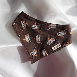 May include: Brown fabric face mask with a pattern of ducks. The mask has a checkered background and a small tag that reads "with care by care". The ducks are in various poses with blue, orange, and black details.