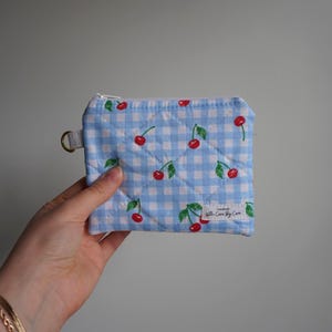 May include: A small, quilted coin purse with a light blue and white checkered pattern. The purse is decorated with red cherries and green leaves. It has a silver zipper and a small metal ring. The text "With Care By Cara" is printed on a white label.