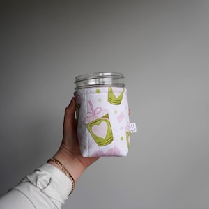 May include: A white jar cozy with a repeating pattern of green drink cups with pink hearts, pink bows, and pink clouds. The cozy is wrapped around a clear glass jar with a silver rim. A small white tag is attached to the side.