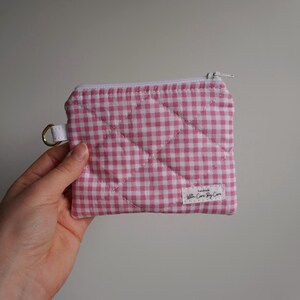 May include: A small, quilted pouch with a pink and white gingham pattern. The pouch has a white zipper, a gold-colored ring, and a small white tag that reads "With Care By Care".