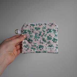 May include: A small, quilted floral coin purse with a white background and a pattern of pink and teal flowers. The purse has a white zipper, a small white strap with a gold-colored D-ring, and the text "with care by care".
