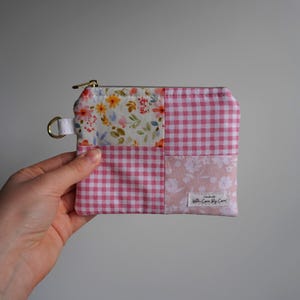 May include: A patchwork zippered pouch with a gold-tone zipper and a small D-ring. The pouch features four fabric squares: floral, pink gingham, and two floral-patterned pink fabrics. The label reads "handmade with Care By Care."