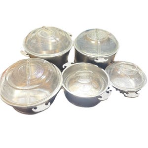 May include: A set of five vintage metal serving bowls with clear glass lids. The bowls are silver-toned with decorative handles and lids. The lids have etched designs. The bowls are of varying sizes, suitable for serving food.