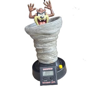 Könnte beinhalten: Eine digitale Looney Tunes Tasmanian Devil Uhr mit einer braunen Figur, die aus einem grauen, wirbelnden tornado-förmigen Sockel ragt. Der schwarze Kunststoffsockel enthält eine Digitalanzeige, einen gelben Knopf und den Text Looney Tunes Tasmanian Devil.