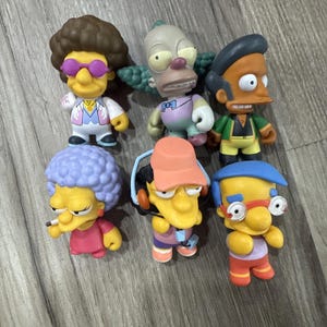 May include: A collection of six Simpsons character figurines. The figurines include Sideshow Bob, Krusty the Clown, Apu Nahasapeemapetilon, Selma Bouvier, Otto Mann, and Milhouse Van Houten. Each figure is brightly colored and detailed.