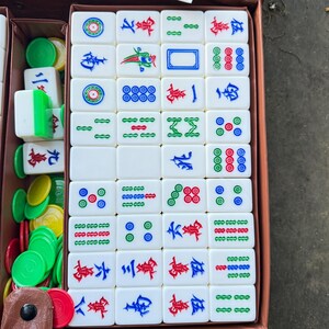 Puede incluir: Un conjunto de fichas de mahjong rectangulares blancas con s&iacute;mbolos grabados en tinta roja, azul y verde, dispuestas en una cuadr&iacute;cula dentro de una caja marr&oacute;n. El set incluye fichas de juego redondas de pl&aacute;stico en colores verde, amarillo y rojo.