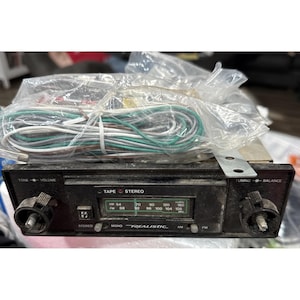 Puede incluir: Una radio de coche Realistic vintage con una placa frontal negra. Cuenta con perillas para tono, volumen, sintonización y balance. La radio tiene configuraciones AM y FM, y un reproductor de cintas. Se incluyen cables y un soporte de montaje.