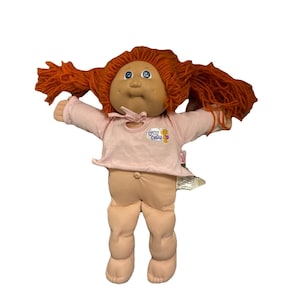 May include: A vintage Cabbage Patch Kid doll with orange yarn hair and blue eyes. The doll wears a light pink shirt with the text "Chou Chou" and a matching diaper. The doll's arms are outstretched.