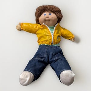 May include: A vintage doll with brown hair, blue eyes, and freckles. The doll is wearing a yellow jacket, a striped shirt, blue jeans, and white socks. The doll's arms are outstretched.