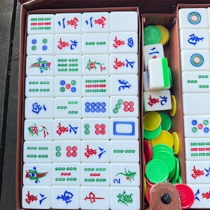 Puede incluir: Un juego de mahjong con fichas rectangulares blancas grabadas con caracteres chinos, patrones de bamb&uacute; y s&iacute;mbolos circulares en rojo, verde y azul. El conjunto incluye una colecci&oacute;n de fichas de puntuaci&oacute;n peque&ntilde;as, redondas y coloridas.