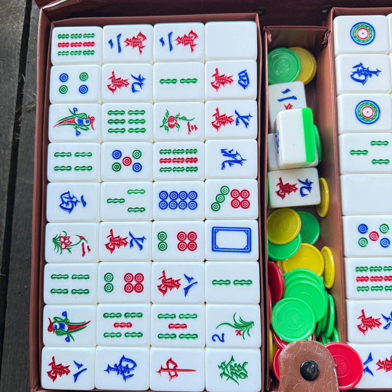 Puede incluir: Un juego de mahjong con fichas rectangulares blancas grabadas con caracteres chinos, patrones de bamb&uacute; y s&iacute;mbolos circulares en rojo, verde y azul. El conjunto incluye una colecci&oacute;n de fichas de puntuaci&oacute;n peque&ntilde;as, redondas y coloridas.