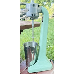 Op de afbeelding: Vintage mintgroene milkshake maker met een roestvrijstalen mengbeker. De mixer heeft een zilveren motorhuis en een gebogen arm. De basis is breed en stabiel. De mixer staat op een houten tafel.