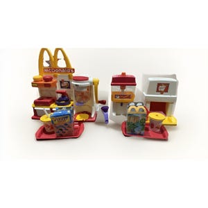 May include: A colorful McDonald's playset featuring a yellow and red building with the McDonald's logo, a drink dispenser, and various food items. Includes trays, cups, and boxes. A fun toy for imaginative play.