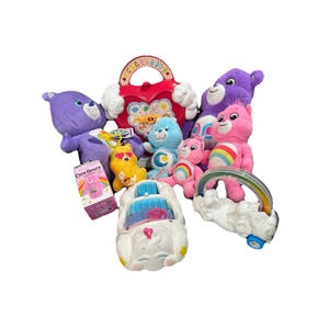 May include: A selection of Care Bears plush toys and accessories in shades of purple, pink, blue, and yellow. The toys include a rainbow-shaped toy, a heart-shaped toy, and a white car-shaped toy.