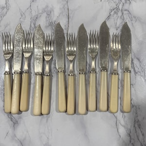 Puede incluir: Un juego de cuchillos de mantequilla y tenedores antiguos. Los cuchillos tienen una hoja curva y mangos de color crema. Los tenedores tienen tres púas y combinan con los mangos de los cuchillos. La cubertería está dispuesta sobre una superficie de mármol.