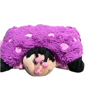 May include: A plush ladybug-shaped pillow in shades of purple, pink, black, and beige. The pillow features a fluffy, textured body with pink polka dots. The ladybug's face has a smiling expression, with black antennae and pink accents.