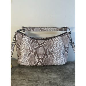 May include: A shoulder bag with a snakeskin pattern in shades of gray and brown. The bag has a zipper closure and a detachable shoulder strap. The hardware is silver-toned. The bag is sitting on a wooden surface.