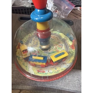 Puede incluir: Un juguete de peonza de estilo vintage con una cúpula transparente que alberga un tren en miniatura sobre una vía circular. El mango central está compuesto por segmentos de plástico apilados en rojo, azul y amarillo. La base es roja.