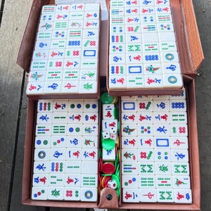 Puede incluir: Un juego de mahjong con fichas rectangulares blancas que presentan caracteres chinos, bamb&uacute; y patrones circulares grabados en color. El set incluye dados blancos peque&ntilde;os y fichas de apuesta redondas de pl&aacute;stico verde y rojo, organizadas en un estuche marr&oacute;n.