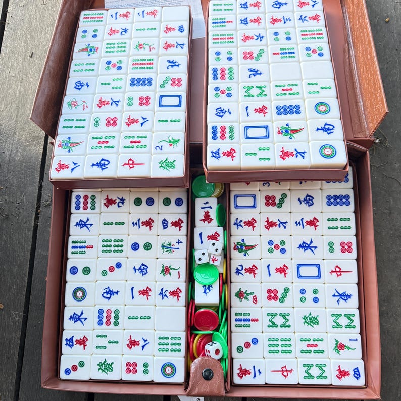 Puede incluir: Un juego de mahjong con fichas rectangulares blancas que presentan caracteres chinos, bamb&uacute; y patrones circulares grabados en color. El set incluye dados blancos peque&ntilde;os y fichas de apuesta redondas de pl&aacute;stico verde y rojo, organizadas en un estuche marr&oacute;n.