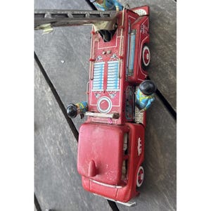 May include: A vintage red tin toy fire truck featuring a long extendable ladder, white and blue side detailing, and two small painted figures in blue uniforms positioned on the sides. The truck has black wheels and a prominent red cab section.