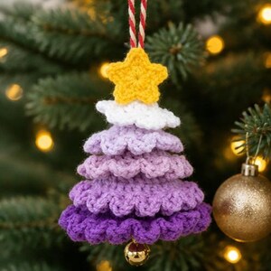 Op de afbeelding: Een gehaakte kerstboomversiering met een gele ster aan de bovenkant, gevolgd door lagen van wit, lichtpaars, middenpaars en donkerpaars garen. Het ornament hangt aan een rood-wit gedraaid koord en is voorzien van een klein gouden belletje.