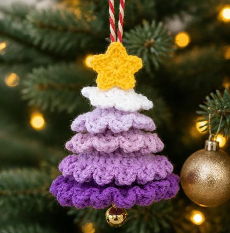 Op de afbeelding: Een gehaakte kerstboomversiering met een gele ster aan de bovenkant, gevolgd door lagen van wit, lichtpaars, middenpaars en donkerpaars garen. Het ornament hangt aan een rood-wit gedraaid koord en is voorzien van een klein gouden belletje.