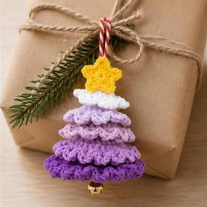 Op de afbeelding: Een gehaakte kerstboomversiering met een gele ster aan de bovenkant, een witte laag en drie lagen paars garen in verschillende tinten. Het ornament hangt aan een rood-wit gestreept koord en heeft onderaan een klein gouden belletje.