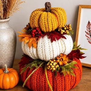 May include: A decorative stack of three crocheted pumpkins in mustard yellow, cream, and burnt orange. The pumpkins are adorned with artificial autumn foliage, including red and orange maple leaves, small yellow berries, and red berries.