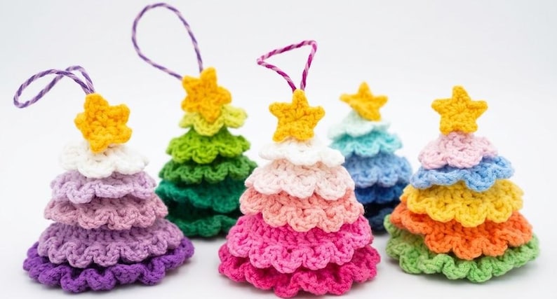 Op de afbeelding: Een verzameling van vijf kleine, kegelvormige gehaakte ornamenten in diverse kleuren. Elk ornament heeft gelaagde, geplooide lagen en is bekroond met een kleine gele ster. Drie ornamenten zijn voorzien van paars-witte gestreepte ophanglussen.