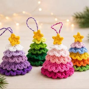 Op de afbeelding: Vier kegelvormige gehaakte ornamenten in de vorm van kerstbomen. Elke boom heeft gelaagde geschulpte lagen in verschillende kleuren, waaronder paars, groen, roze en regenboogkleuren, bekroond met een kleine gele ster en een paars-witte lus.