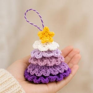 Op de afbeelding: Een klein, kegelvormig gehaakt ornament met een verloop van paarse lagen, bekroond met een witte laag en een gele ster. Het ornament is voorzien van een paars en wit gedraaid koordje aan de bovenkant om het op te kunnen hangen.