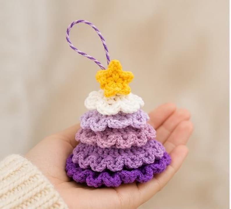 Op de afbeelding: Een klein, kegelvormig gehaakt ornament met een verloop van paarse lagen, bekroond met een witte laag en een gele ster. Het ornament is voorzien van een paars en wit gedraaid koordje aan de bovenkant om het op te kunnen hangen.