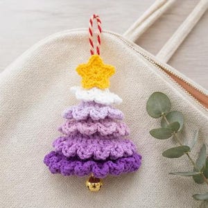 Op de afbeelding: Een gehaakte kerstboomhanger met een gele ster aan de bovenkant, gevolgd door lagen van wit, lichtpaars, middenpaars en donkerpaars garen. Een klein goudkleurig belletje hangt aan de onderkant, met een rood-wit gedraaid koordje als lus.