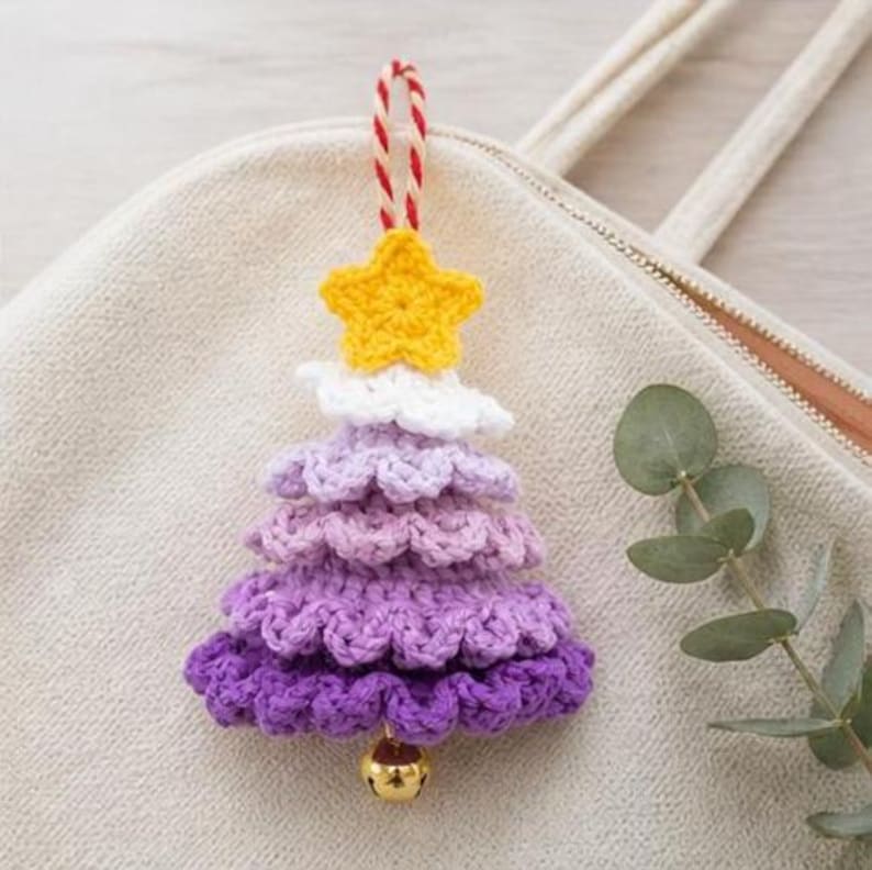 Op de afbeelding: Een gehaakte kerstboomhanger met een gele ster aan de bovenkant, gevolgd door lagen van wit, lichtpaars, middenpaars en donkerpaars garen. Een klein goudkleurig belletje hangt aan de onderkant, met een rood-wit gedraaid koordje als lus.