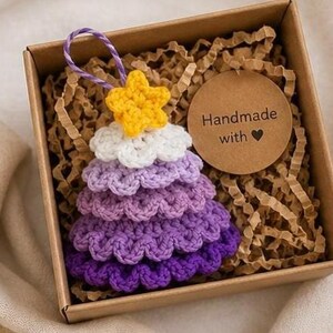 Op de afbeelding: Een gehaakte kerstboomversiering met lagen garen in een paars kleurverloop, bekroond met een gele ster en een paars-wit ophangkoordje. Het ornament zit in een bruine kartonnen doos met een rond label met de tekst Handmade with.