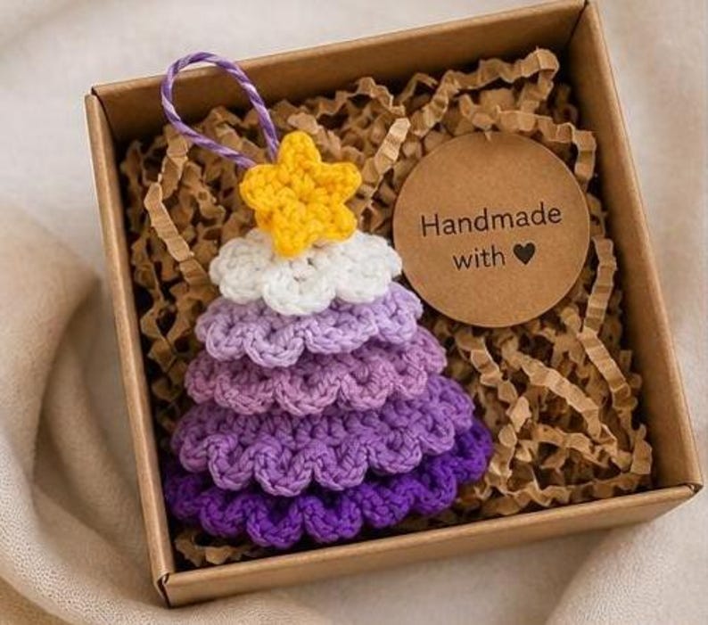 Op de afbeelding: Een gehaakte kerstboomversiering met lagen garen in een paars kleurverloop, bekroond met een gele ster en een paars-wit ophangkoordje. Het ornament zit in een bruine kartonnen doos met een rond label met de tekst Handmade with.