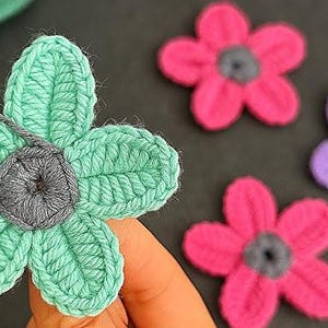 May include: Close-up of handmade crochet flowers in various colors. One flower is held by a hand, showing a mint green petal design with a gray center. Other flowers are pink and purple, also with gray centers. The flowers are on a dark surface.