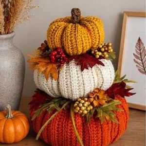 May include: A stack of three crocheted pumpkins in mustard yellow, cream, and burnt orange. The pumpkins are decorated with artificial autumn leaves, red berries, and small gold-toned spheres nestled between the layers of textured yarn.