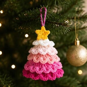 Op de afbeelding: Een gehaakte kerstboomversiering met een verloop van roze en witte garenlagen, bekroond met een klein geel gehaakt sterretje. Het ornament hangt aan een roze en wit gedraaid koordje en heeft een getextureerd ontwerp met verschillende lagen.