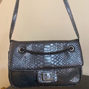 May include: A dark grey shoulder bag with a textured snakeskin pattern. The bag features a rectangular flap, a metallic turn-lock closure, and a decorative dark metal chain detail across the front. It includes a matching shoulder strap.