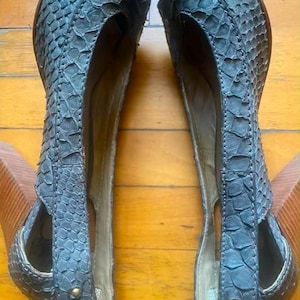May include: A pair of dark grey shoes with a textured snakeskin pattern, featuring cut-out sides, a rounded toe, and a wooden block heel. Each shoe is accented with a small, circular brass-toned metal stud near the heel and the toe area.